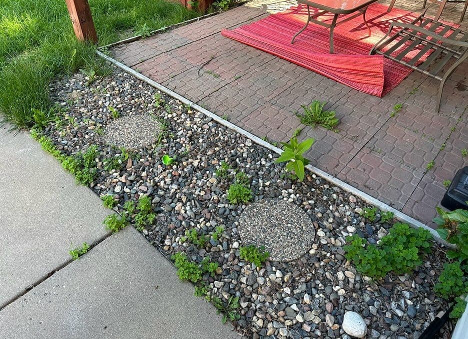 Patio area with stone borders, gravel, green plants, and a red outdoor rug beside furniture.