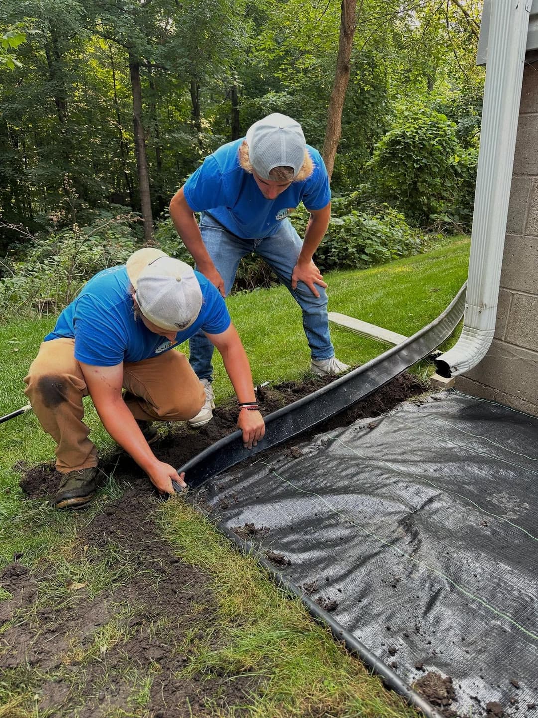 Two workers installing drainage around a house, using tools and materials on a grassy area.