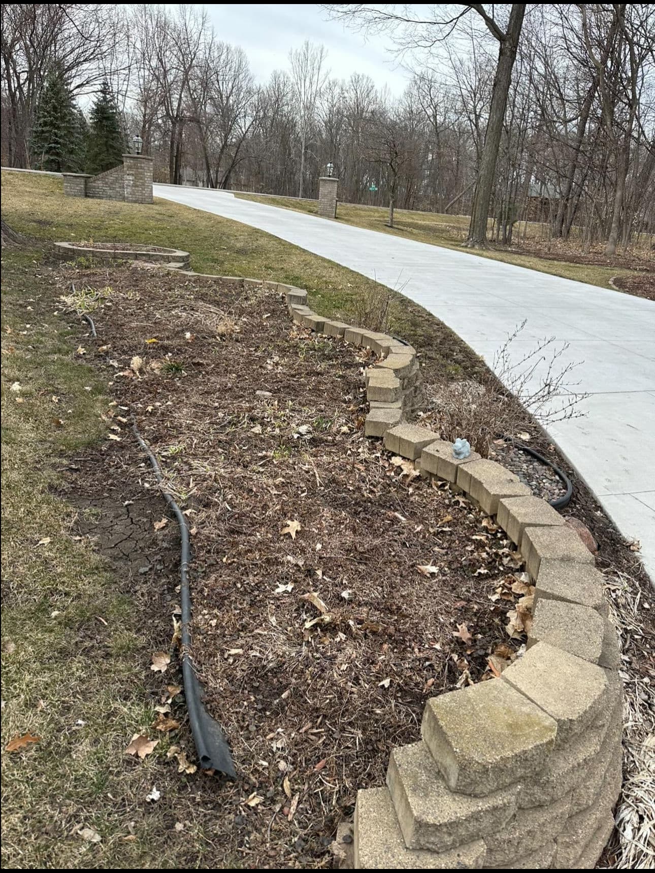 Bare garden bed with stone border alongside a paved driveway and trees in the background.