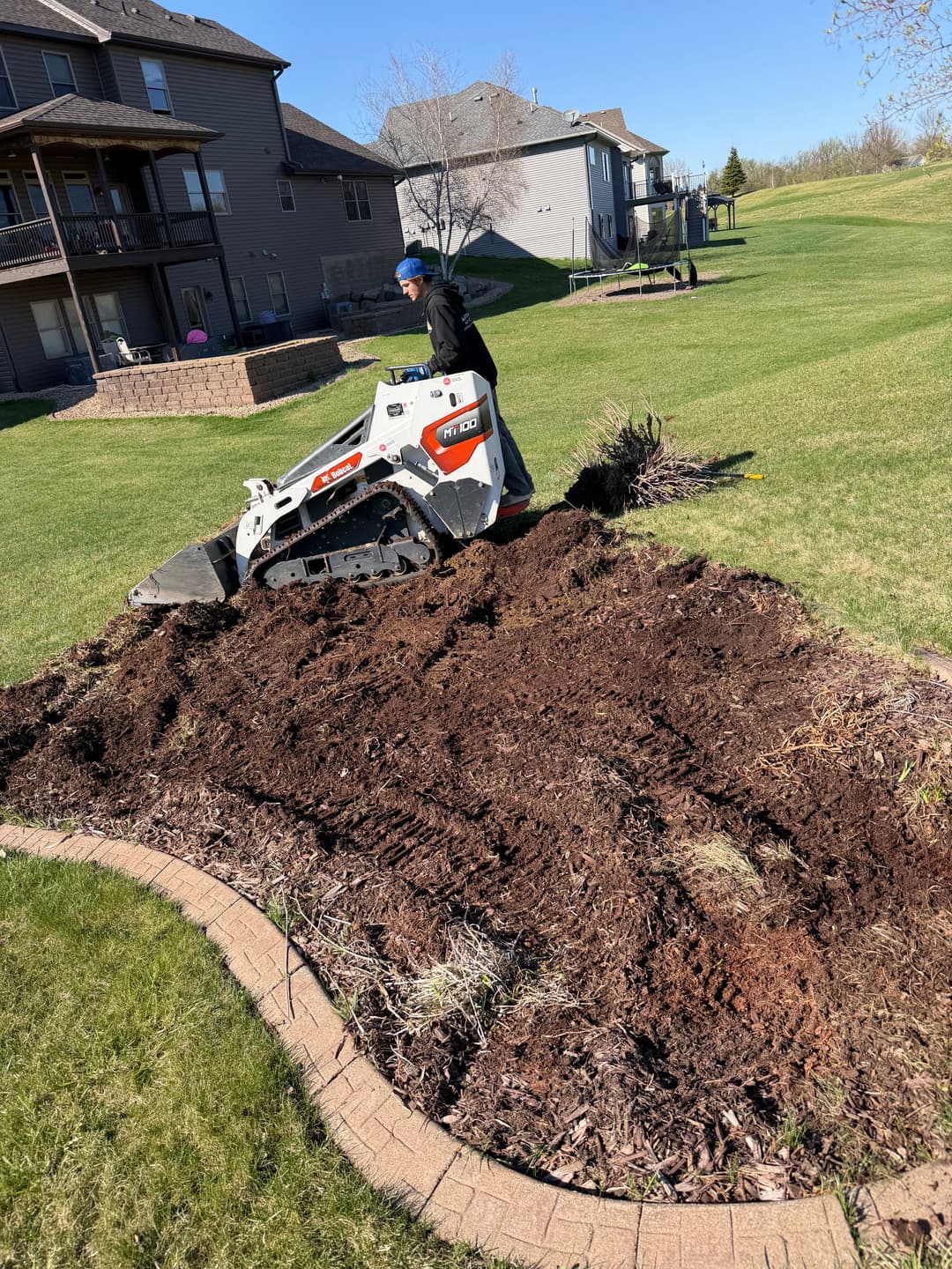 Man using a compact track loader to landscape a yard, with mulch and homes in the background.