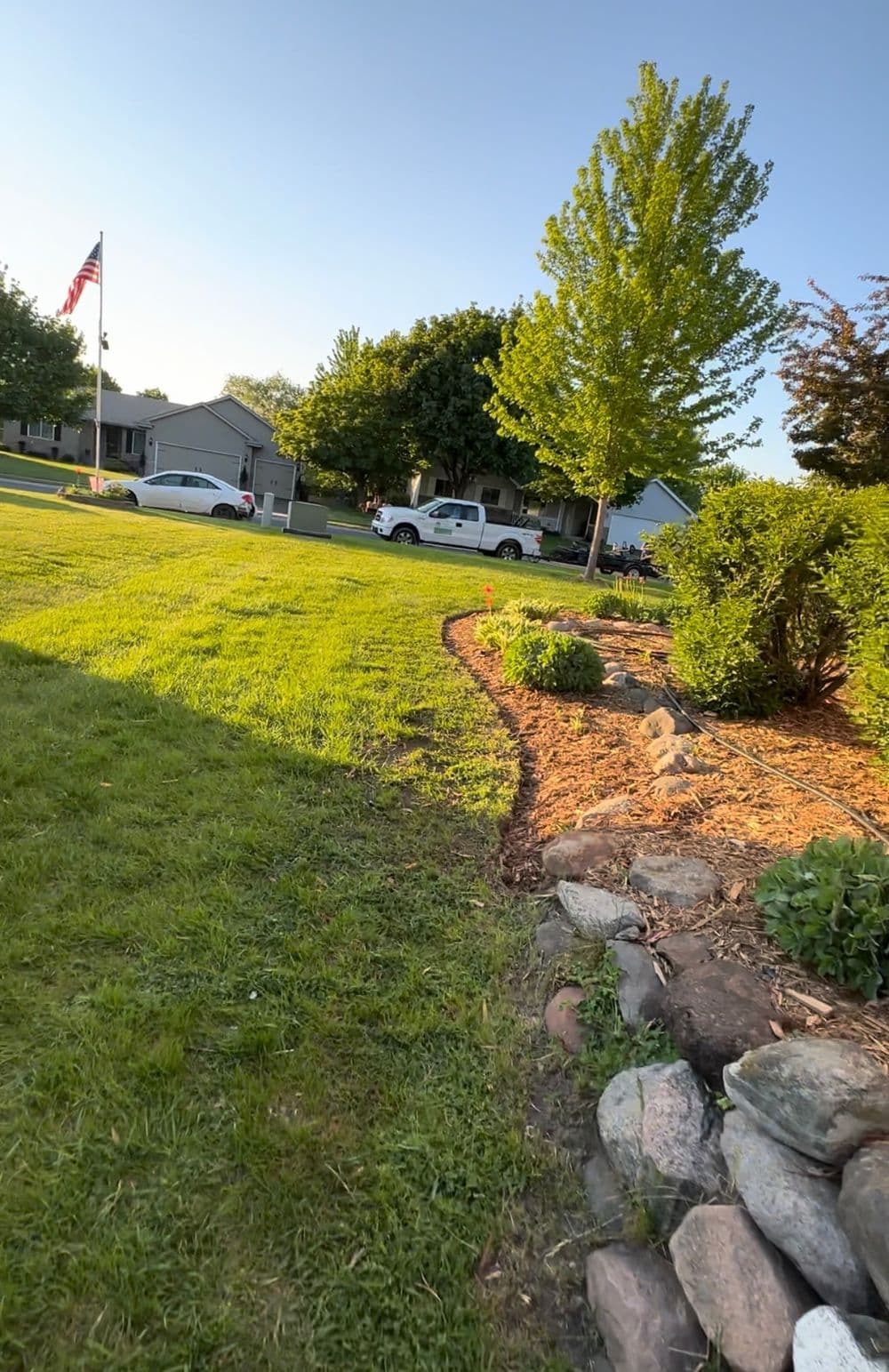 Lush green lawn with garden beds, American flag, and houses in suburban neighborhood.