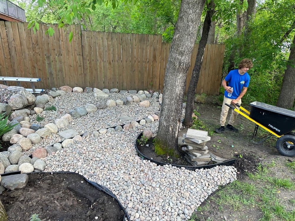 Teenager landscaping a backyard with gravel pathway and stone features near trees.