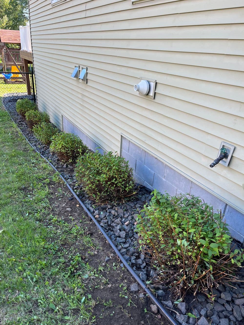 Bushes along the side of a house with gravel landscaping and utility connections.
