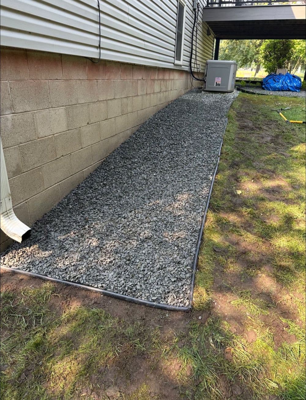 Gravel pathway alongside a house, leading to an air conditioning unit and landscaped area.